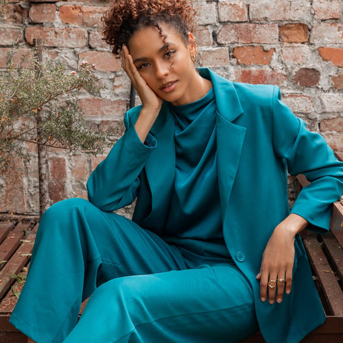 A woman wearing a matching teal blue  two-piece suit with a structured blazer and wide-leg trousers, seated on a wooden bench against a rustic brick wall.