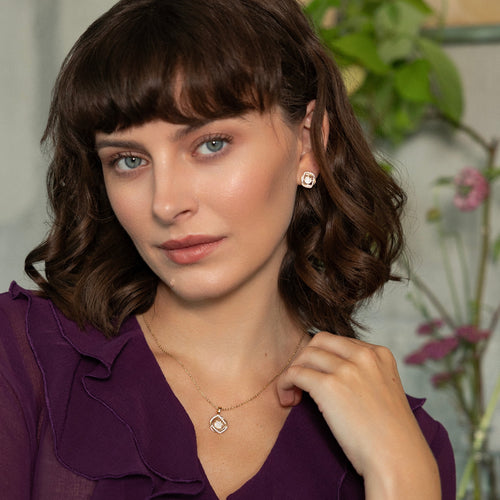 A woman with brown hair and bangs wearing a purple blouse, a delicate gold necklace with a square pendant, and a matching stud earring, set against a softly blurred indoor background with green plants.