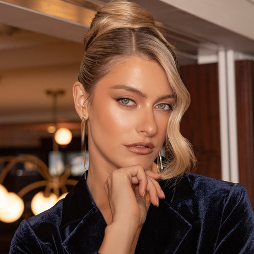 A close-up portrait of a woman with blonde hair styled in a half-up bun, wearing a dark navy velvet blazer and long gold hoop earrings, set against a softly blurred indoor background with warm lights.