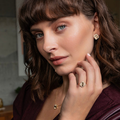 A close-up lifestyle shot of a woman with brown hair and blue eyes, wearing a gold ring and matching gold necklace with heart-shaped charms, and a gold flower-shaped earring. She is wearing a dark plum top against a softly blurred indoor background.