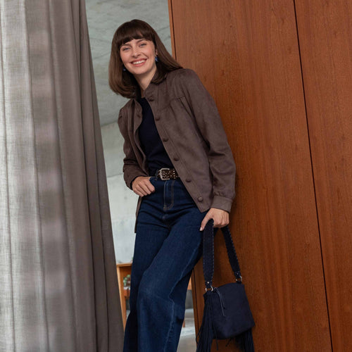 A woman stands indoors wearing a brown button-front overshirt over a black top and dark blue jeans, holding a dark blue handbag. She is posed in front of a large wood-paneled wall with a curtain on the left.