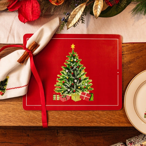 A red rectangular Christmas-themed placemat featuring an embroidered-style green Christmas tree with presents, styled on a wooden table with a white napkin tied with a red ribbon and holiday greenery.