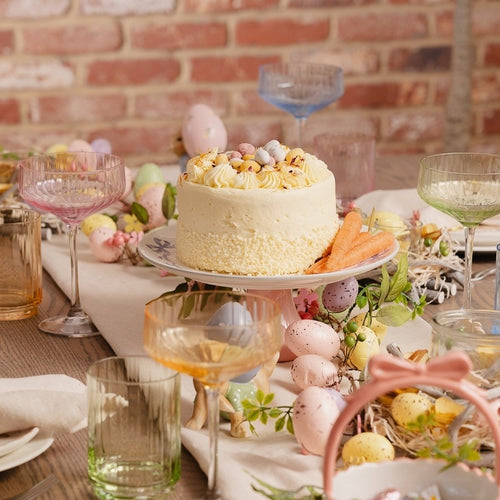 Easter-themed table setting with a cake, eggs, and decorative glasses against a brick wall.