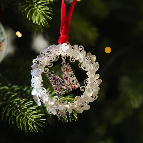 A silver-toned metal Christmas ornament shaped like a wreath with a hanging red ribbon and a central letter N embellished with small multicolored rhinestones, shown hanging on a dark green Christmas tree branch.