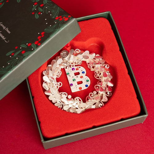 A silver-toned monogram letter B ornament decorated with colorful rhinestones, nestled in a red velvet-lined gift box next to its dark green holly-patterned lid on a red background.