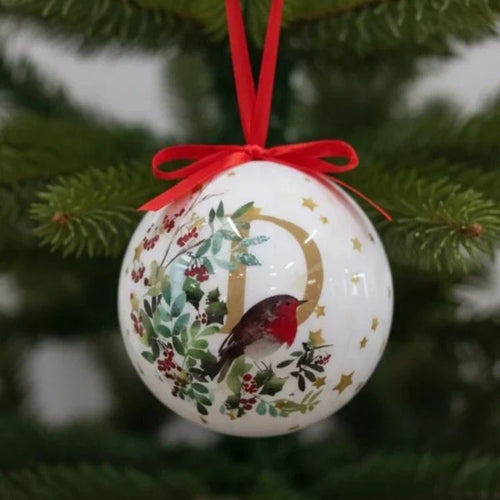 A white Christmas ornament shaped like a ball, decorated with a gold letter D, a robin bird, holly leaves, red berries, and small gold stars. It is hung by a red ribbon on a green Christmas tree branch.
