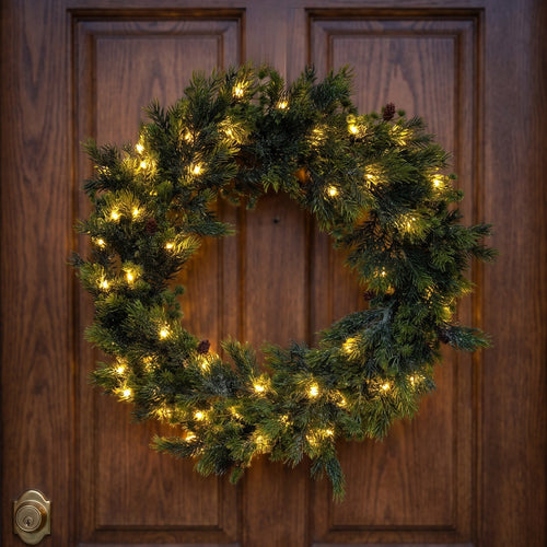 A green artificial Christmas wreath with warm white LED lights and small pinecones, hanging on a dark brown wooden door.