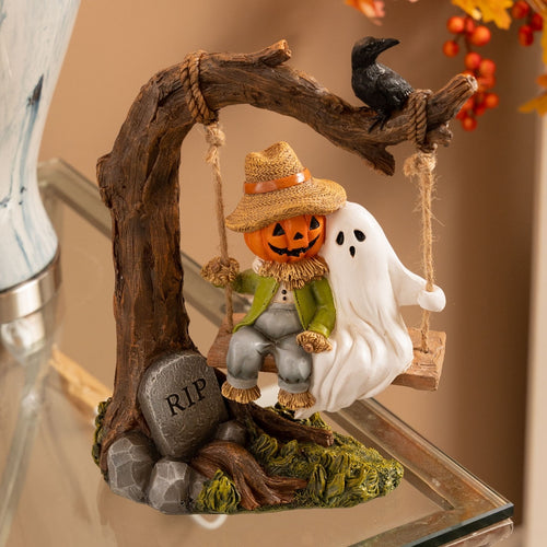 Halloween-themed resin figurine featuring a scarecrow on a swing with a pumpkin head, a white ghost, a black raven perched on the branch, and a small RIP stone at the base, displayed on a glass shelf.