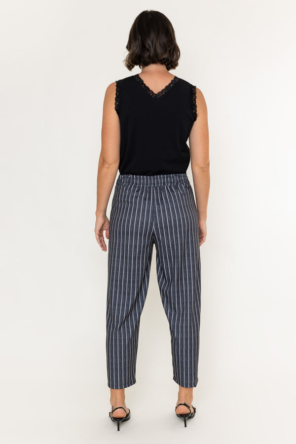 A woman viewed from the back wearing a black sleeveless top with lace trim and high-waisted navy trousers with thin white vertical stripes, paired with black heels, against a plain white studio background.