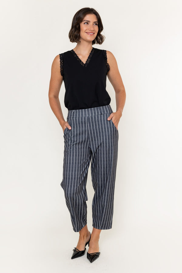 A woman modeling a sleeveless black top with lace trim and high-waisted navy blue pinstriped trousers, paired with black pointed-toe heels, against a plain white studio background.