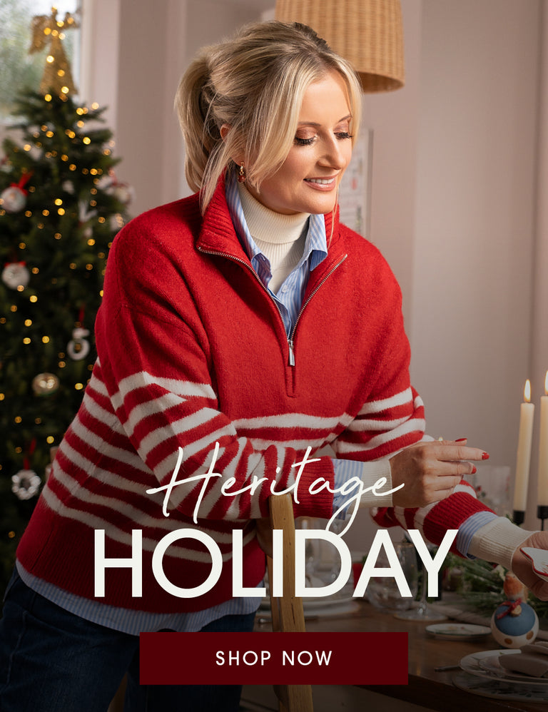 A woman in a cozy indoor setting wearing a red and white striped quarter-zip sweater over a white turtleneck and blue shirt. A decorated Christmas tree is visible in the background, and large text overlays read 'Heritage HOLIDAY' with a 'SHOP NOW' button at the bottom.