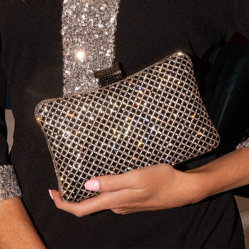 A close-up of a person holding a sparkling rhinestone-covered clutch bag. The person is wearing a black dress with silver embellishments, and the clutch features a grid pattern of clear crystals that reflect light.