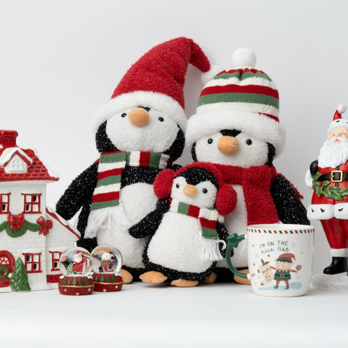 Three penguin plush toys wearing festive Christmas hats and scarves, arranged with a small house, snow globes, a mug, and a Santa figurine on a white studio background.