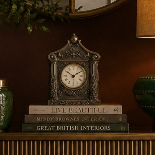 An ornate antique-style metal table clock with a white clock face and black Roman numerals, displayed on top of three decorative books on a wooden mantelpiece in a warm, dark interior setting.