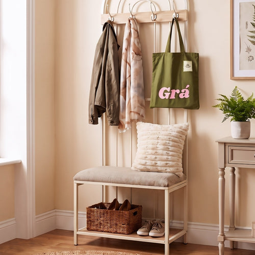 A cream-colored wooden entryway bench with a cushioned seat and open lower shelf, styled in a warm beige hallway. A green tote bag with pink text reading 'Grá' hangs on the back rack, along with a jacket and scarf.