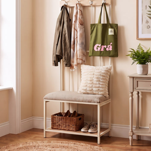 A cream-colored entryway bench with a cushioned seat and open lower shelf, styled in a warm beige room. A green tote bag with pink text, a brown jacket, and a scarf hang from hooks on the back, with a textured cream pillow resting on the seat and shoes in the basket.