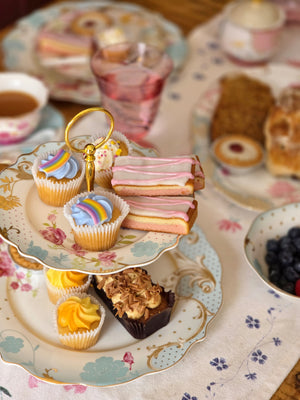 A styled afternoon tea spread on a floral porcelain table setting, featuring a two-tiered stand with cupcakes, pastries, and sandwich cookies, alongside teacups and a glass of pink drink on a wooden table.