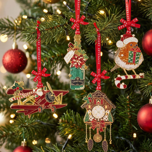 Four Christmas tree ornaments hanging on a decorated Christmas tree with warm lights: a red biplane, a green bottle ornament with gold lettering, a cuckoo clock, and a gingerbread bird with a Santa hat and gift.