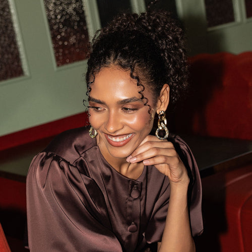 A woman with curly dark hair wearing large gold and silver hoop earrings with clear stones, posing in a warm indoor setting with a red chair and green wall in the background.