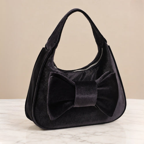 A black velvet shoulder bag with a large bow on the front, set on a light marble surface against a beige background.