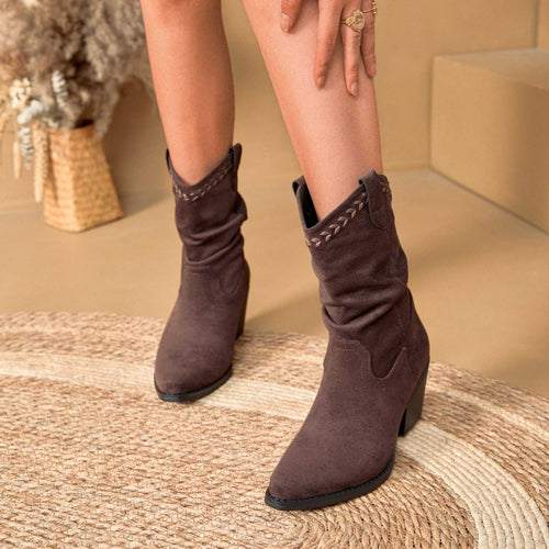 A person wearing dark brown suede ankle boots with a slouchy fit, decorative stitching, and a low block heel, standing on a woven rug in a warm beige indoor setting.