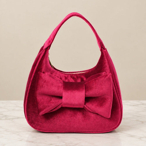 A bright fuchsia velvet shoulder bag with a large matching bow on the front, set against a neutral beige background.