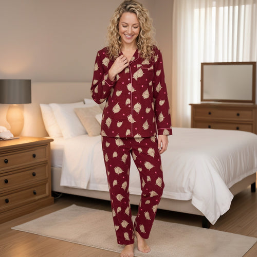 A woman stands in a bedroom wearing a matching long-sleeve pajama set with a burgundy base and beige tree pattern. The set includes a button-front top and matching pants.