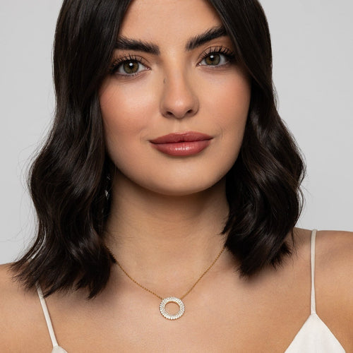A close-up of a woman with dark wavy hair wearing a delicate gold necklace with a circular pendant encrusted with small clear stones, set against a plain light gray background.