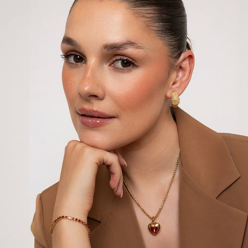 A close-up lifestyle shot of a woman wearing a gold heart pendant necklace with a red heart-shaped stone, a matching gold earring, and a gold bracelet, styled with a brown blazer against a light gray background.