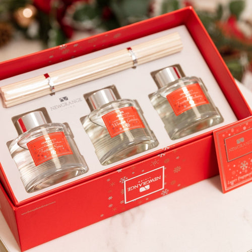 A New Balance fragrance gift set in a red box, featuring three clear glass bottles with silver caps and orange labels, plus a light wood diffuser stick on a white insert.