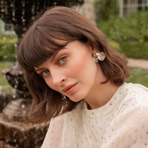 A woman with a brown bob haircut and bangs wearing a white embroidered top and a large flower-shaped earring, photographed outdoors in front of a blurred garden background.