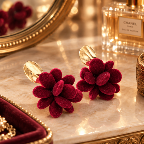 A pair of deep burgundy velvet flower-shaped earrings with gold-tone metal backs, displayed on a polished marble surface next to a Chanel perfume bottle and an ornate gold mirror.