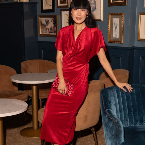 A woman wearing a shiny red wrap-style midi dress with short sleeves and a V-neckline, standing in a furnished lounge with framed wall art, brown chairs, and blue velvet seating.