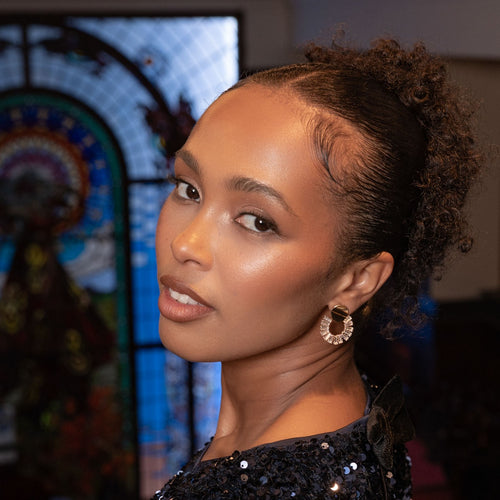 A close-up portrait of a woman with styled curly hair pulled back, wearing a sparkling dark sequined top and a gold-toned earring, set against a softly blurred interior background with a stained-glass window.